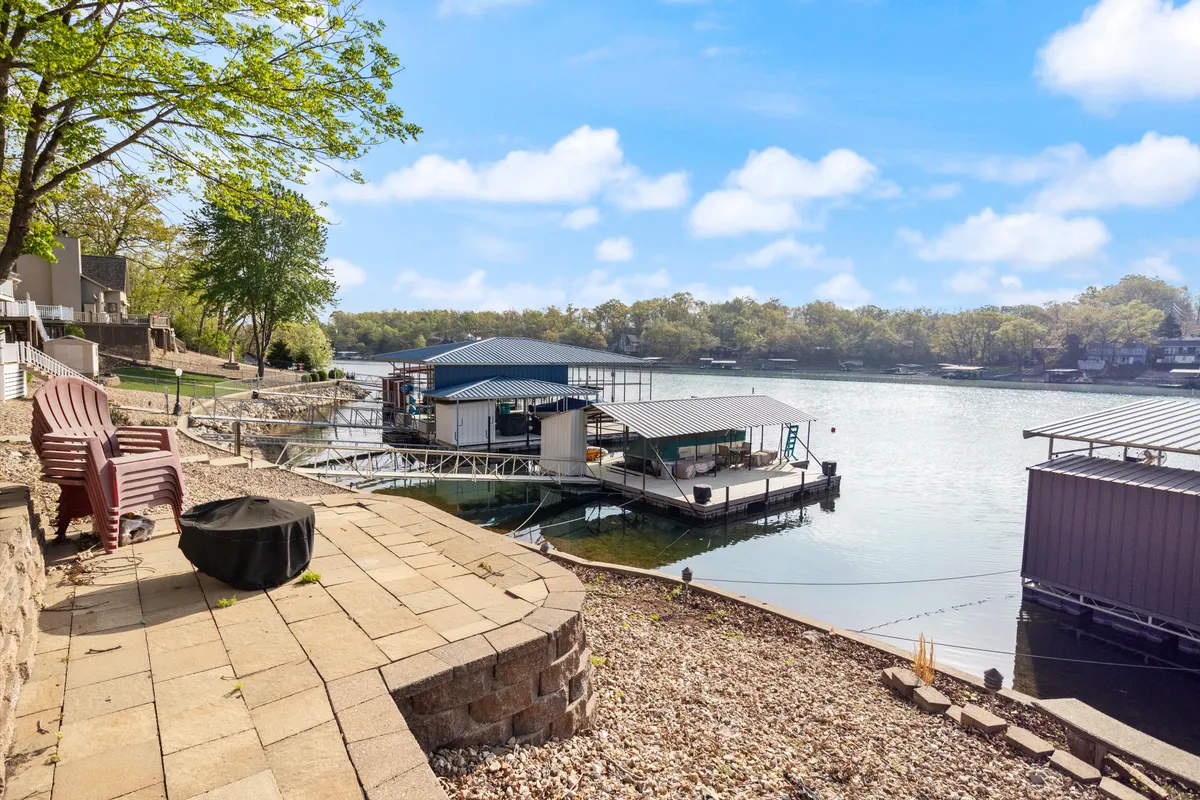 This exterior shot features a serene lakeside setting with a dock, Adirondack chairs, and a fire pit overlooking the calm water. The view highlights morning reflections and the welcoming atmosphere of the lakefront, inviting guests to launch watercraft, gather, or simply relax by the shore.