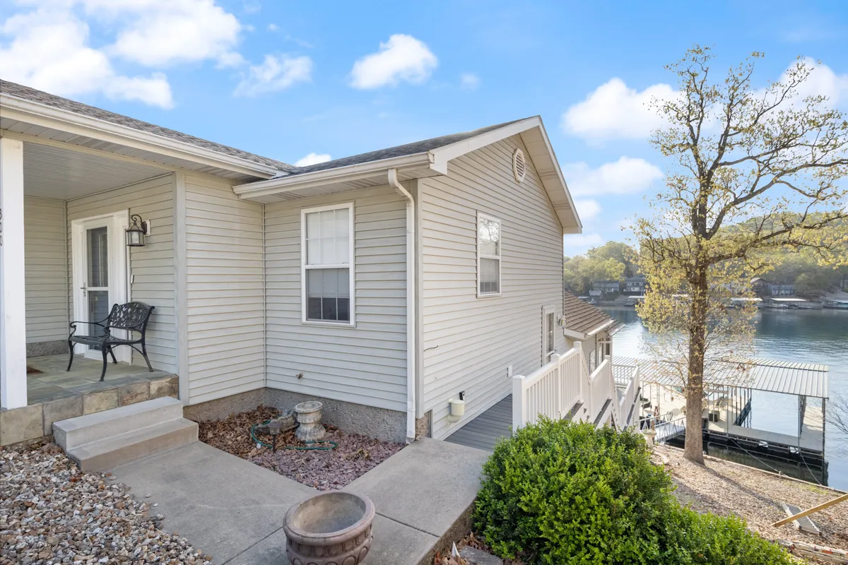 A welcoming front porch overlooks the lake, with lush bushes framing the view and a scenic pathway leading up to the home. The lake glimmers in the background, suggesting tranquil mornings and beautiful sunset vistas. The porch is an inviting spot to relax regardless of the season.