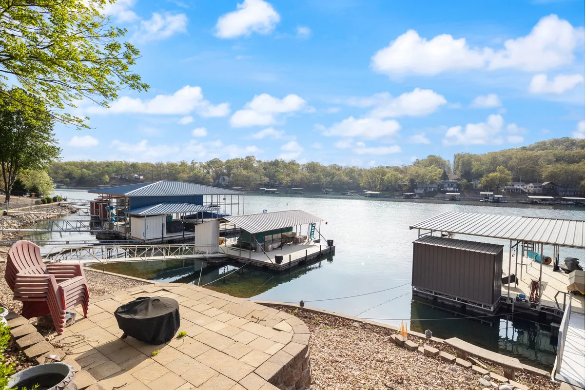 An exterior view featuring a patio with comfortable seating, a scenic lake with distant boathouses, and a tree-lined backdrop. The inviting setup suggests relaxation, lakeside dining, and group gatherings as the sun dips toward the horizon.