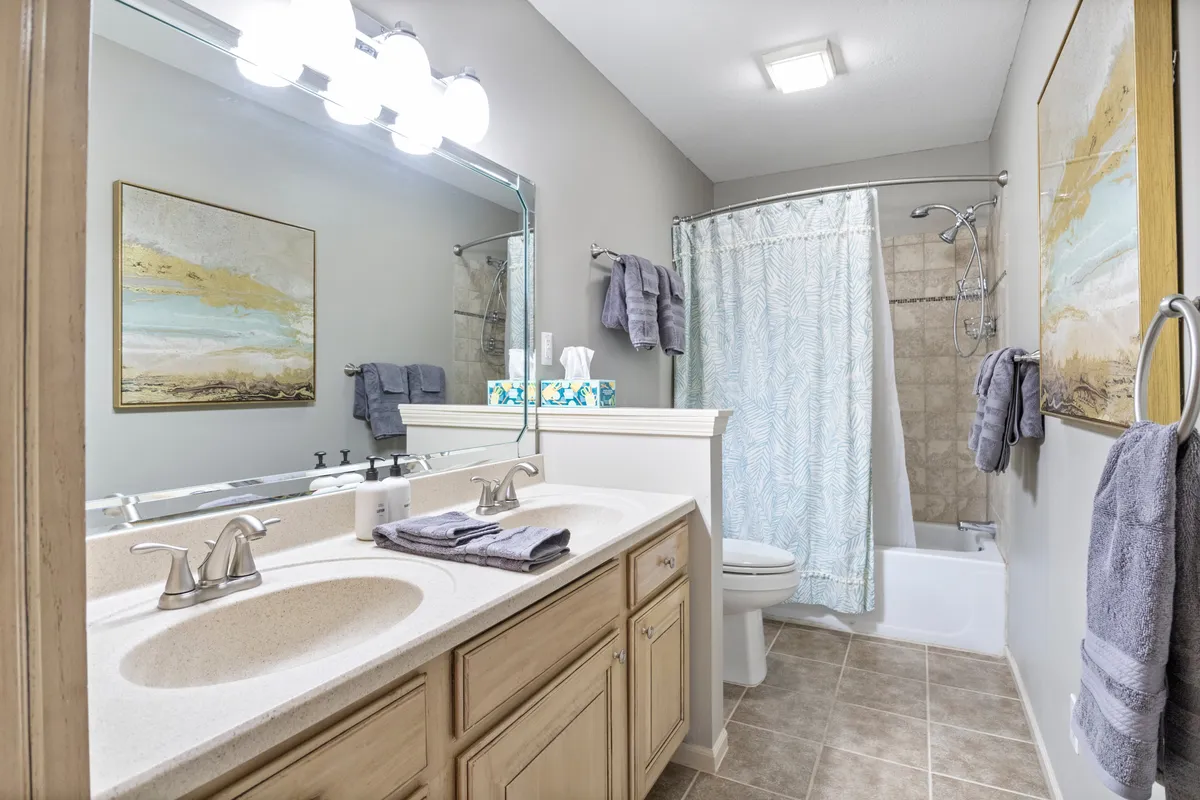 A modern bathroom with double sinks, a large mirror, a relaxing bathtub, and tasteful artwork. The space is refreshing and serene, suggesting a high standard of care and thoughtful amenities.