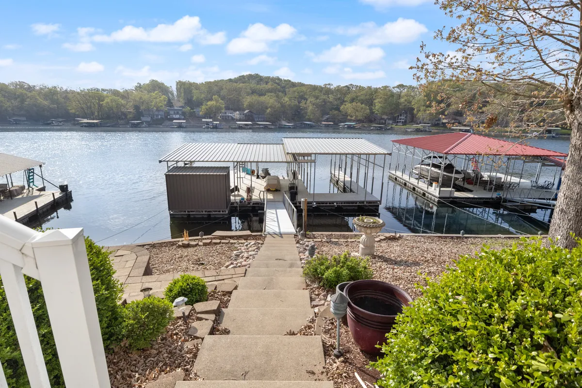 A spacious dock extends into the lake, surrounded by lush trees and featuring steps down to the water. Boathouses can be seen in the background, and the dock area includes places to sit and enjoy the view.