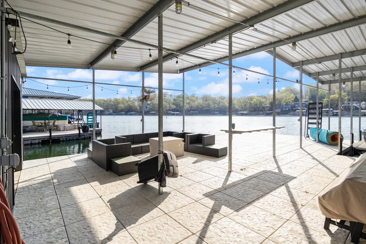 A spacious dock lounge with comfortable seating overlooks a calm lake at sunset. Decorative string lights add ambiance, while a boat dock and water view in the background create a serene, inviting atmosphere perfect for relaxing and socializing.