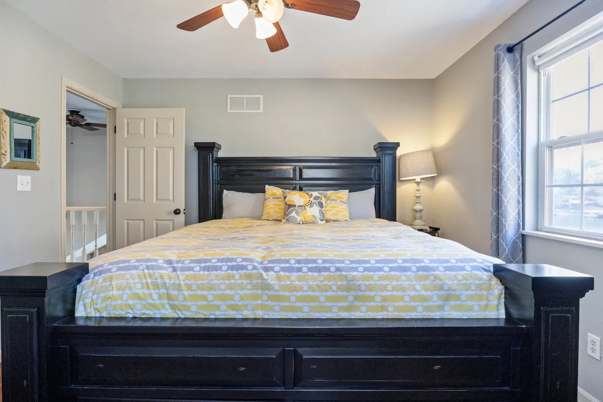 A spacious bedroom featuring a large bed with patterned bedding, a decorative lamp, a window that lets in natural light, and a framed mirror. The room is styled for comfort with tranquil decor and ample space for relaxation or reading.