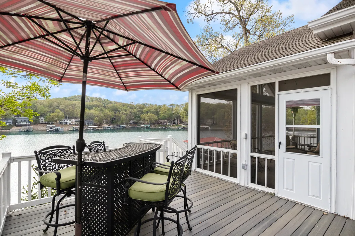 A serene outdoor dining table with a striped umbrella sits on a deck overlooking the lake and tree-lined shore. The setting feels tranquil and inviting, ideal for enjoying meals, coffee, or cocktails while soaking in the view.