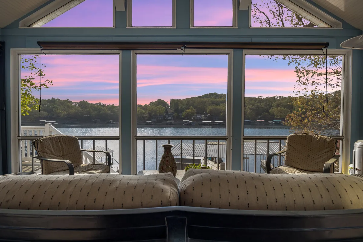 The image showcases a sunlit living area with plush, comfortable seating arranged to take in expansive lake views through floor-to-ceiling windows. A decorative vase adds a touch of elegance. The space is bright, serene, and effortlessly inviting, with the tranquil lake and vibrant sunset visible just outside.