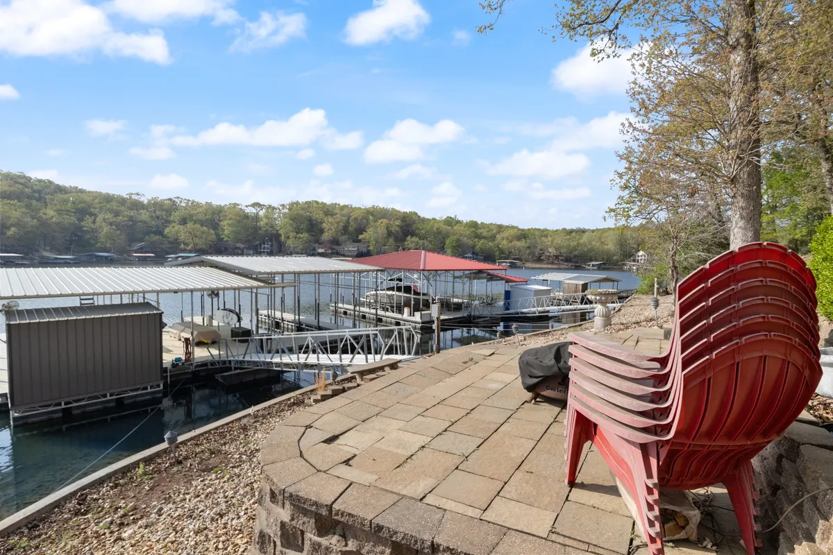Red chairs are gathered on a patio by the water, with a boathouse and tree line in the background. The setting evokes a sense of peaceful gathering—ideal for morning coffee, sunset viewing, or evening bonfires by the lake.