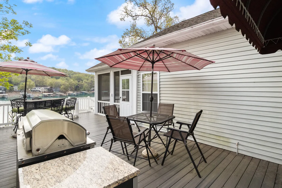 This image captures an expansive outdoor deck with a grill, dining table, umbrellas, and a beautiful lake view. The space is set up for both dining and lounging, with the lake shimmering in the background. The casual arrangement feels ideal for social gatherings, meals outdoors, and taking in the scenery.
